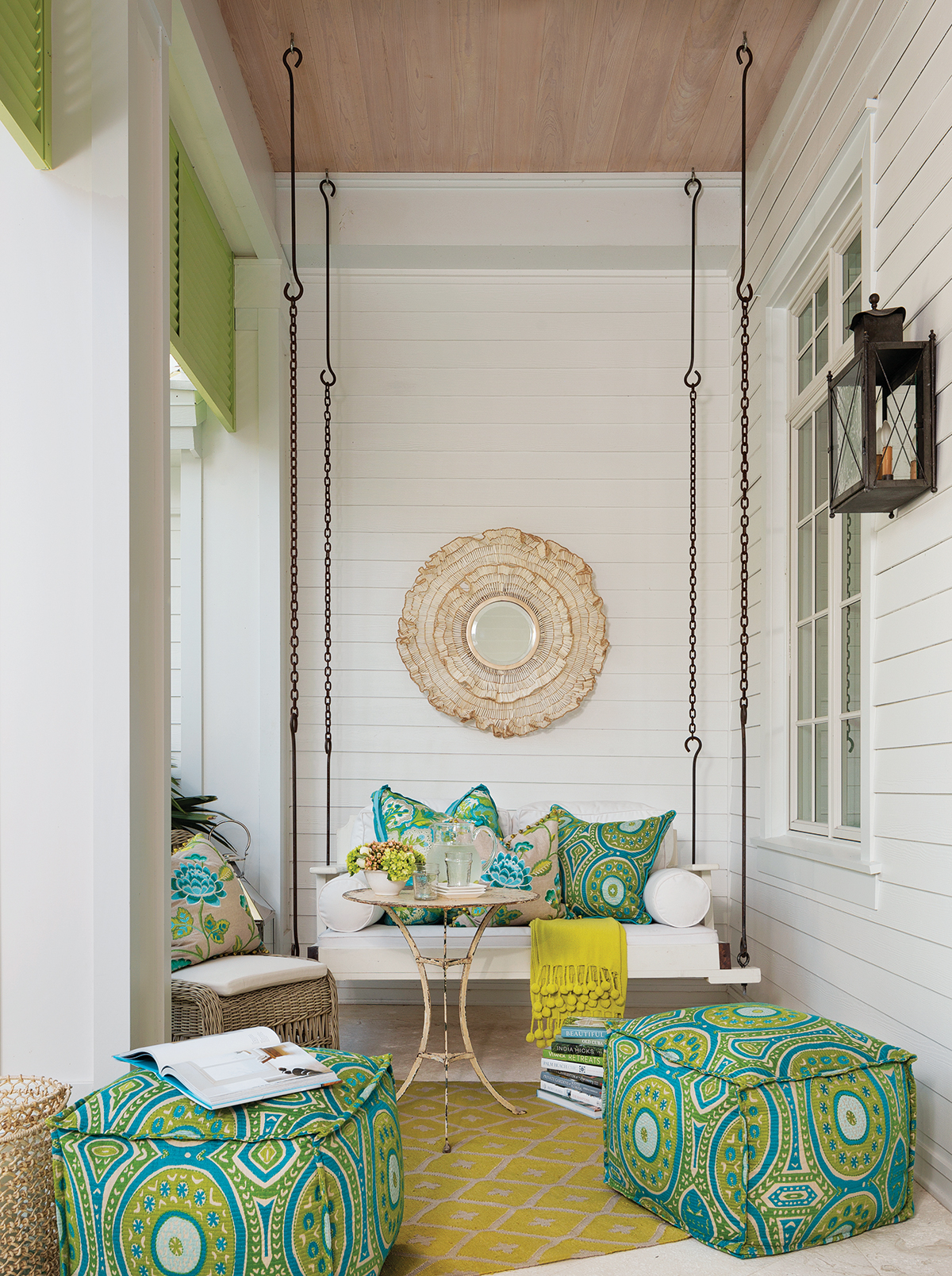 Bright patterned fabrics in ottomans and accent pillows continue outdoors on the cozy front porch. A rattan mirror from Palecek and a vintage bistro table convey an earthy vibe, while ottomans clad in a chartreuse-and-turquoise print provide extra seating.