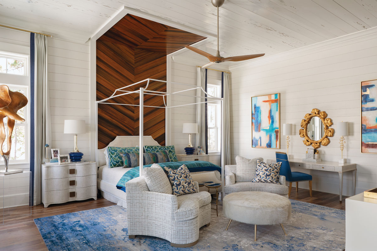 A mahogany chevron wall accent wraps the master bedroom’s Old Biscayne canopy bed in warmth. The curvilinear lines of the club chairs, nightstands, and ottoman soften the hard finishes. Paintings by artist Tyler Arnold flank a vanity table.