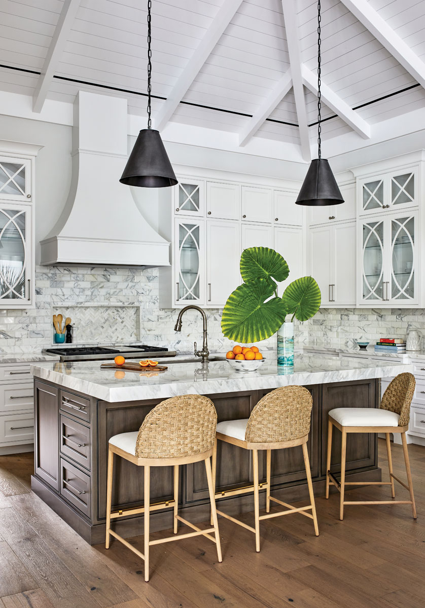 : The island countertop in the kitchen is clad in Calacatta ORO marble. Tucked beneath the lip that extends beyond the dark cabinetry are counter stools made of oak with woven backs and linen seats. The pendants hanging above are made of black glass.