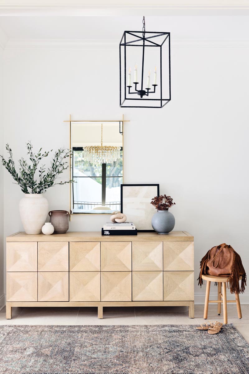 Interior designer Julianne Hendrickson updated the home’s original front entrance with a delightfully simple and streamlined Noir console table and mirror. A patterned area rug brings texture to the foyer’s clean aesthetic. A single wrought-iron chandelier overhead lends a touch of elegance.