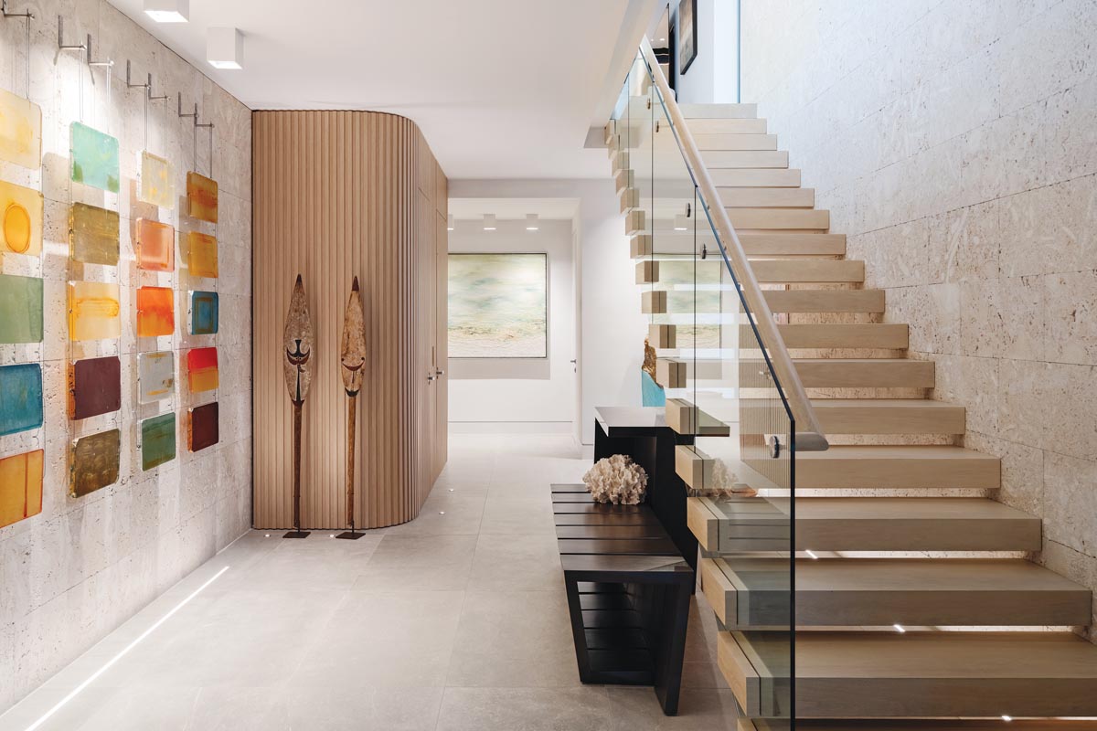 On the first floor foyer, Five Hanging Ladders, a colored glass installation by Carrie McGee, is backdropped by the same coral stone that’s used in the home’s exterior. The ribbed oak enclosure to the left of the stairwell conceals the elevator that connects the residence’s three levels.