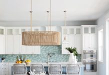The kitchen emanates a chic nautical vibe, bringing ocean hues into the heart of the home. A glass-tile backsplash mixing clear tones of seafoam green and aqua backdrops a brilliant blue island that anchors the space. Natural fiber accents like rope barstools from Essentials For Living and the dining table’s linear Currey & Company rope chandelier create textural interest.