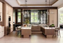 The kitchen’s twin islands appear to float atop walnut-stained cerused oak and plinth supports clad in textured copper. Visual Comfort lighting fixtures illuminate the marble surfaces, which are heavily veined in a deep wine hue. Fluted tambour wood panels trim the perimeter.