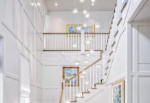 The beach-inspired foyer sets a serene tone with sandy white oak floors, a sky-inspired Stark stair runner, and a two-story beaded chandelier from Currey & Company. White paneled walls and a barrel-vaulted ceiling enhance impressionist paintings by local artist Janine Wesselmann.