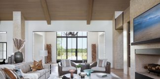 The living room’s double-sided fireplace—shared with an adjacent club room—generates a sculptural centerpiece, lending warmth and ambiance while maintaining an open-concept layout. A sizable sofa from Lazar Industries and cozy Century club chairs establish a conversation area around a trio of marble-topped Palecek cocktail tables.