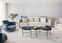 The living room’s coastal-chic aesthetic is anchored by a plush, textured rug from Tai Ping Carpets. A cream-colored Adriana Hoyos sofa is adorned with blue pillows, mirroring the deep navy tones of the private elevator foyer. Olivya Stone swivel armchairs and a trio of nested Jerry Pair cocktail tables reiterate the inky blue hue.