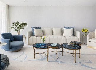 The living room’s coastal-chic aesthetic is anchored by a plush, textured rug from Tai Ping Carpets. A cream-colored Adriana Hoyos sofa is adorned with blue pillows, mirroring the deep navy tones of the private elevator foyer. Olivya Stone swivel armchairs and a trio of nested Jerry Pair cocktail tables reiterate the inky blue hue.