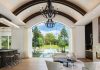 Custom Elegance in Estates at Bay Colony Golf Club Home Bent walnut radius beams sweep across the ceiling, concealing air systems and allowing the architecture by John Cooney to read as clean, continuous, and intentional. Chandeliers are wrapped in black leather, introducing a tactile contrast that softens the scale. Integrated lighting is tucked seamlessly into the structure, eliminating visual clutter and keeping the focus on form and material rather than mechanical elements. The effect draws the eye forward to the dramatic window at the back of the great room, engineered as a single uninterrupted plane of glass.